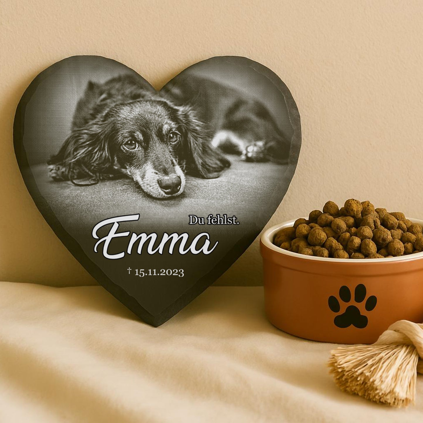 Herzförmiger Gedenkstein mit Foto eines liegenden Hundes, Text 'Emma Du fehlst. † 15.11.2023', daneben Napf mit Pfotenmotiv und Hundetrockenfutter