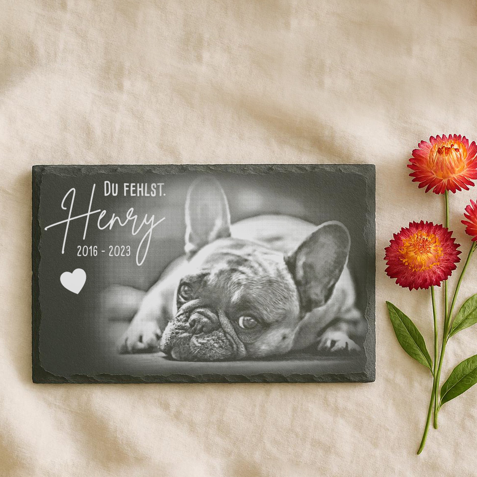 Granitplatte mit schwarz-weißer Abbildung eines französischen Bulldogs, Text 'Du fehlst. Henry 2016 - 2023' und Herzsymbol. Neben einer roten Blume.